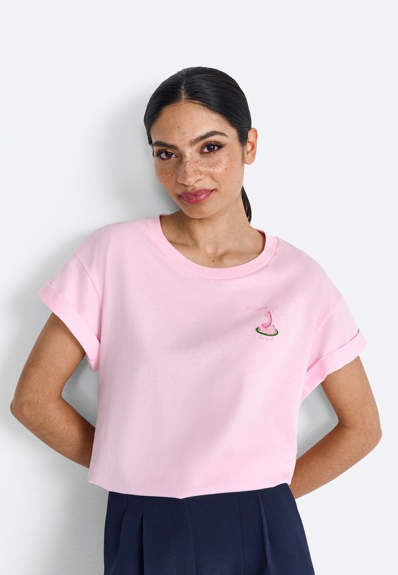 Roze katoenen T-shirt met korte opgerolde mouwen, voorzien van een klein geborduurd ontwerp op de borst. Korte lengte, zachte textuur.