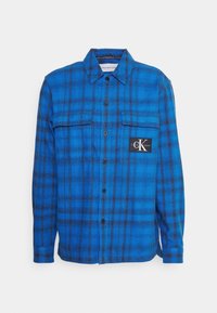 Blue and black plaid flannel shirt with long sleeves, front buttons, two chest pockets, and a black Calvin Klein logo patch.