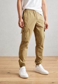 Pantalons cargo beige avec poches latérales, taille élastique, poignets fuselés et texture douce, associés à des baskets blanches sur un sol en bois.