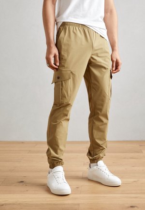 Beige Cargo-Hosen mit seitlichen Taschen, elastischem Bund, schmal zulaufenden Bündchen und einer glatten Textur, kombiniert mit weißen Turnschuhen auf einem Holzboden.