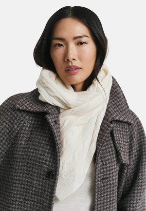Vrouw in een witte kabelgebreide sjaal en een bruine houthakkerswollen jas, recht vooruit kijkend met een neutrale uitdrukking tegen een witte achtergrond.