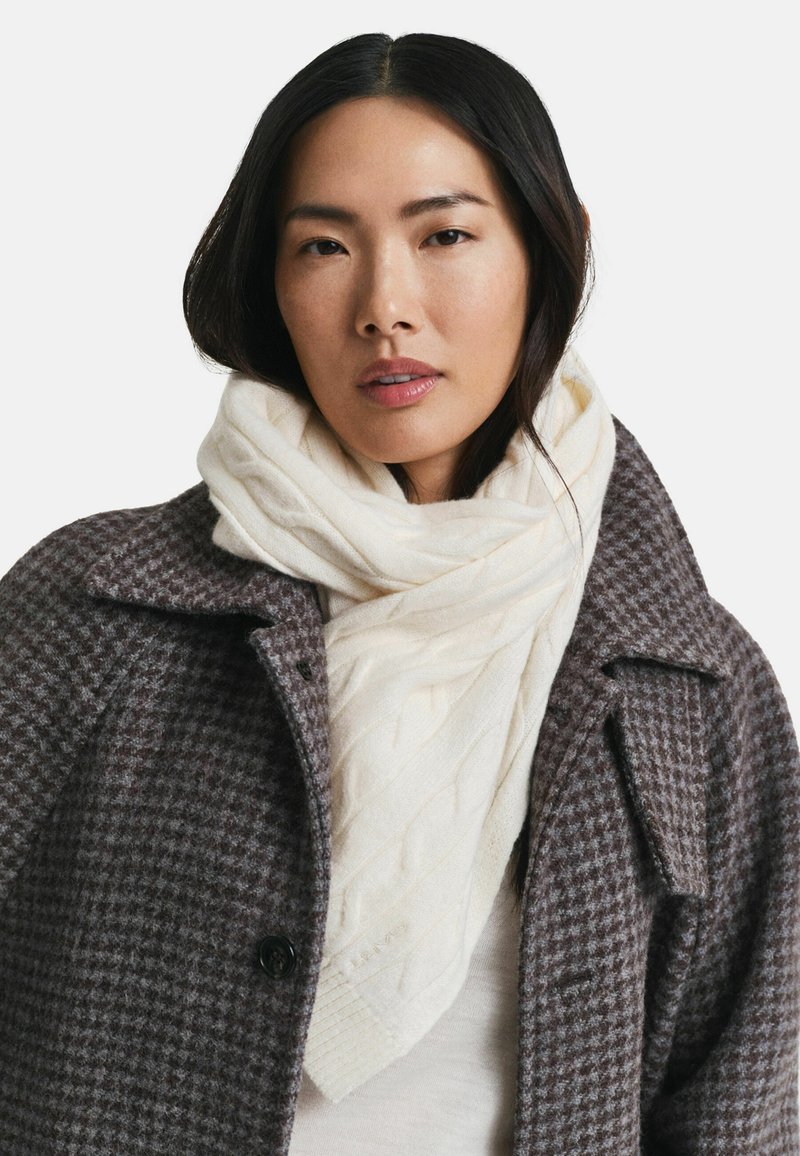 Femme portant une écharpe blanche en tricot à torsades et un manteau en laine à motif pied-de-poule marron, faisant face à l'avant avec une expression neutre sur un fond blanc.