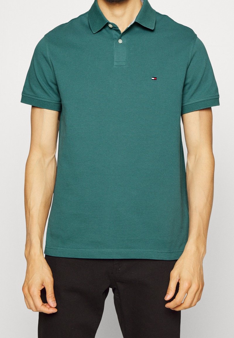 Homme portant un polo vert à manches courtes avec un petit logo Tommy Hilfiger sur la poitrine gauche et un pantalon noir.