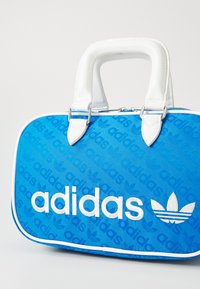 Blå stoffhåndveske med hvite detaljer, med preget Adidas-logo og trefoil-design. Runde håndtak og glidelås-lukking.