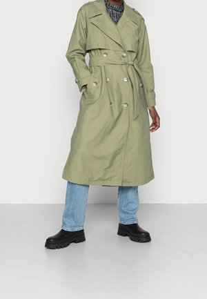 Largo abrigo trench verde oliva con botones cruzados y cinturón, combinado con vaqueros azul claro y botas negras gruesas.