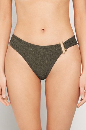 Kobieta w teksturowanych, ciemnozielonych majtkach bikini z prostokątną złotą klamrą na prawym boku, widziana od talii do górnej części ud.