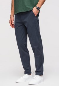 Homme portant un pantalon bleu foncé, des baskets blanches, une chemise verte texturée et une montre en argent au poignet, avec une main dans la poche sur fond gris.