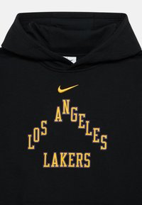 Melns krekliņš ar kapuci no kokvilnas ar priekšā esošu grafiku: "LOS ANGELES LAKERS" dzeltenā un violetā krāsā, virs teksta dzeltenā Nike logo.