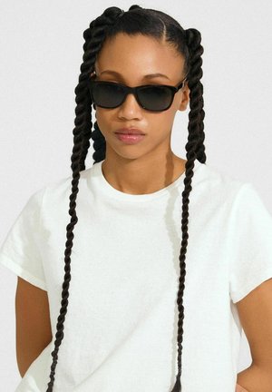 Crne pravokutne sunčane naočale s sjajnim završetkom, uparene s jednostavnom bijelom majicom. Subjekt ima dugačke, isprepletene pramenove koji uokviruju lice.