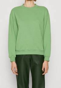 Sudadera de manga larga en algodón verde claro con cuello redondo, corte relajado, puños y dobladillo acanalados; combinada con pantalones verdes oscuros.
