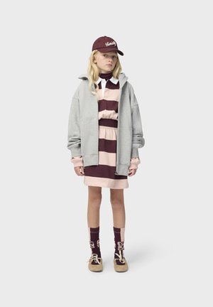 Giovane persona che indossa un vestito a righe color amaranto e rosa, felpa con cappuccio grigio chiaro con cerniera, berretto amaranto, calzini abbinati e scarpe beige, in piedi davanti a uno sfondo uniforme.