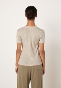 Woman with dark hair in a bun wearing a beige short-sleeve shirt and olive pants, standing facing away against a plain background.