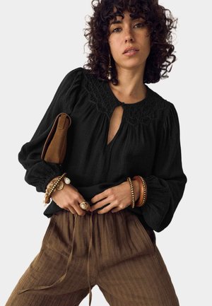 Kvinde med krøllet hår iført sort bluse med lange ærmer, brune teksturerede bukser, guldbelagte og perlearmbånd, en ring, og bærer en brun clutch under armen.