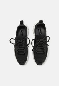 Melni sporta apavi, kas izgatavoti no teksturētas auduma ar plakanām šņorēm. Zole ir balta, ar noapaļotu dizainu un pastiprinātu papēža daļu.