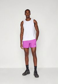 Camisola branca sem mangas, calções roxos com o logótipo da Nike branco, ténis pretos e meias até ao tornozelo, em pé contra um fundo simples.