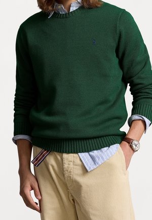 Mężczyzna noszący ciemnozielony dzianinowy sweter na niebiesko-białej pasiastej koszuli, beżowe spodnie sztruksowe oraz zegarek na nadgarstku z skórzanym paskiem.