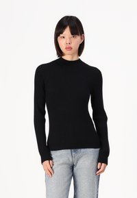 Pull en tricot noir côtelé avec un col montant et des manches longues, associé à un jean délavé clair avec des embellissements subtils.