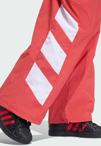 Červené tepláky s bielymi diagonálnymi pruhmi, vyrobené z ľahkej tkaniny. Skombinované s čiernymi teniskami s červenými akcentmi a vzorovanou špičkou.