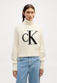 Cremfarbener Rollkragenpullover mit geripptem Muster und schwarzem ‚CK‘-Logo. Kürzes Design mit langen Ärmeln, kombiniert mit hellblauen Jeans.