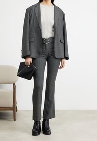 Donna che indossa un blazer grigio a spina di pesce e pantaloni abbinati con cintura, top a costine crema, stivaletti in pelle nera alla caviglia, mentre tiene una borsa nera.