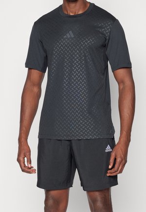 Homme portant un t-shirt de sport Adidas noir avec un motif subtil en forme de losange et un short Adidas noir, sur un fond uni.