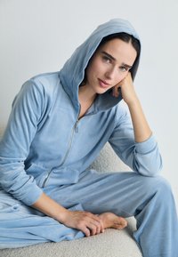 Ljusblå velourjumperset med dragkedja fram, långa ärmar och stor huva. Har en framficka och mjuk textur.