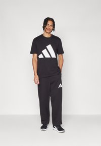 Sort bomulds T-shirt med stort hvidt tre-stribe logo og sorte træningsbukser med et matchende logo. Fodtøj er sort med hvide accenter.