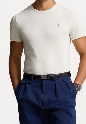 Homme portant un polo blanc à manches courtes, un pantalon bleu marine plissé avec ceinture en cuir, et une montre-bracelet, les mains dans les poches, sur un fond uni.