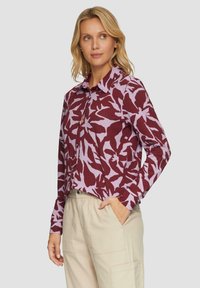Langarmshirt in bordeaux en lavendel met een bloemenpatroon. Voorzien van een kraag en knoopsluiting. Gedragen met lichte broek.