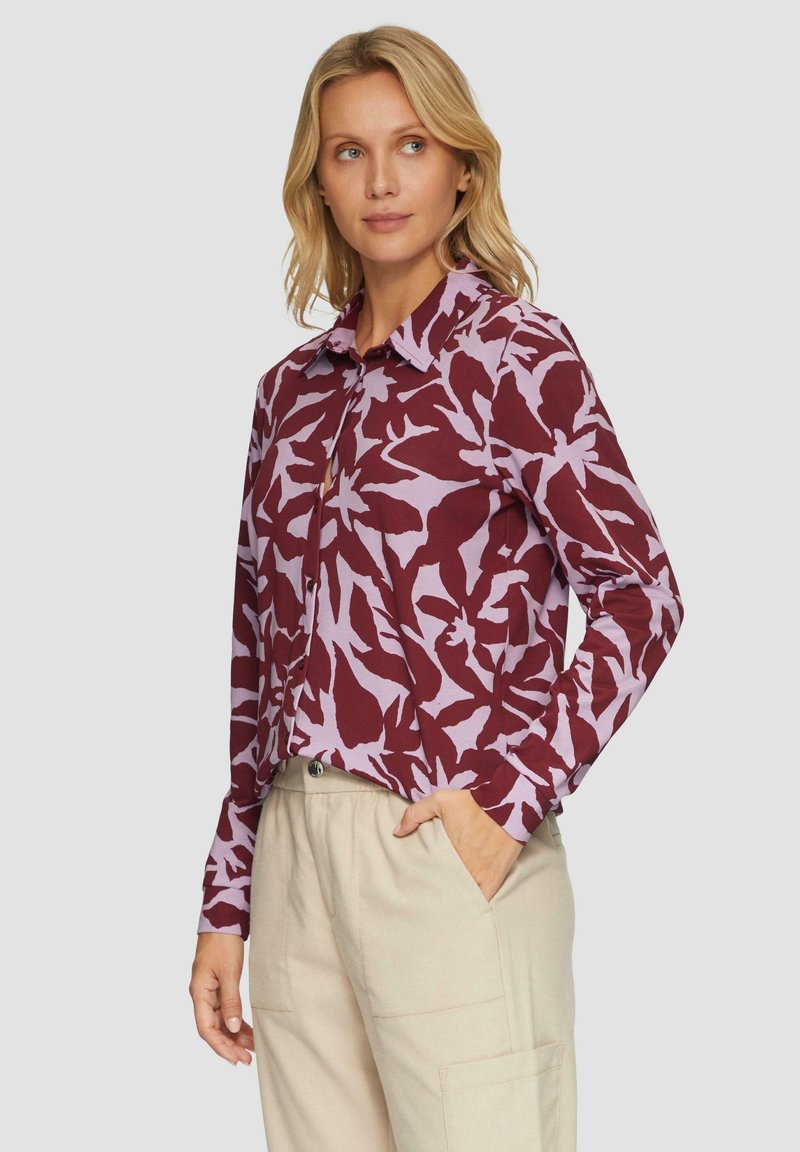 Langarmshirt in bordeaux en lavendel met een bloemenpatroon. Voorzien van een kraag en knoopsluiting. Gedragen met lichte broek.