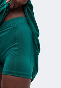 Κοντινό πλάνο ενός ατόμου που σηκώνει ένα πράσινο πουκάμισο, φοράει εφαρμοστά πράσινα αθλητικά σορτς με ορατές ραφές στο πλάι και στη μέση.