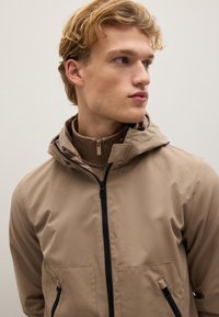 Giacca impermeabile beige con cappuccio, dotata di due tasche frontali, zip nere e una texture liscia. Design leggero per maggiore confort.