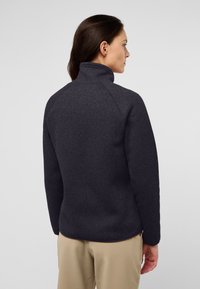 Pullover in pile grigio scuro con colletto alto, tessuto strutturato, maniche raglan e silhouette aderente, abbinato a pantaloni chiari.