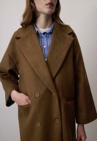 Vrouw in een bruine dubbelknopige jas en een blauw gestreepte overhemden, met een hand in de jaszak.