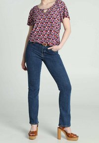 Femme portant un chemisier à manches courtes à motifs colorés, un jean bleu et des sandales marron à talons compensés, debout avec une main dans la poche.