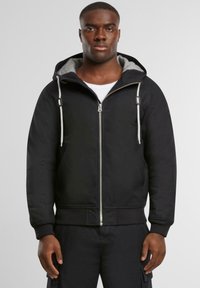 Svart hettegenser med glidelås og gråfôret hette, justerbare snorer og ribbestrikkede mansjetter. Har en jevn tekstur og frontlommer.