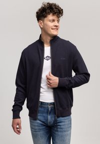 Granatowa kurtka z zamkiem błyskawicznym o teksturowanej powierzchni, z prążkowanymi ściągaczami i białym akcentem logo. Noszona na białej koszulce i zestawiona z jasnoniebieskimi dżinsami.