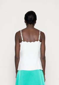 Donna con la pelle scura e i capelli raccolti a chignon, che indossa un top senza maniche con bordo a scallop bianco e una gonna turchese, rivolta di spalle alla fotocamera.