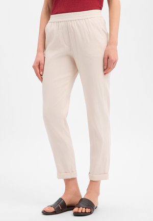 Femme portant un pantalon beige clair retroussé, un haut rouge et des sandales noires à enfiler, debout devant un fond blanc uni.