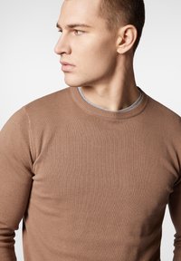 Brązowy sweter dzianinowy z okrągłym dekoltem, teksturowanym materiałem i żebrowanymi mankietami. Modelka pokazana jest w profilu na jasnym tle.