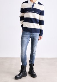 Marinblå och krämrandig rugbytröja med krage och logotyp, kombinerad med ljusblå jeans och svarta snörade kängor. Slät tygtextur.