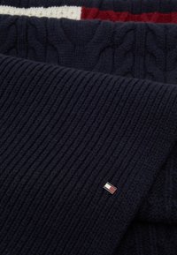 Navy knitted scarf featuring ribbed and cable patterns, complemented by a small metal logo accent with red and white details.