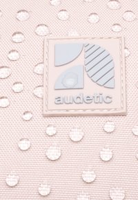 Ružová textúrovaná látka s vyvýšenými priehľadnými kvapkami; obsahuje štvorcové logo v svetlomodrej farbe s geometrickými tvarmi a slovom "audetic" nižšie.