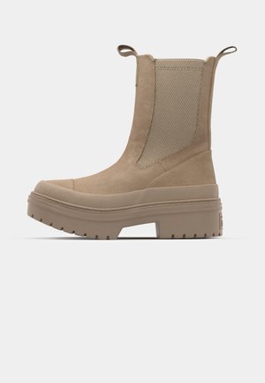 Botín tobillo color tostado con un upper de ante suave, acentos de malla, paneles laterales elásticos y una suela de goma gruesa con detalle de tracción.
