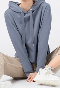 Ljusgrå hoodie med dragsko och metallöglor, kombinerad med beige byxor och klumpiga vita sneakers. Mjukt tyg och avslappnad passform.