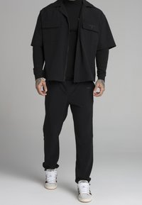 Svart kortärmad zip-up skjorta med två bröstfickor, kombinerad med svarta smala byxor och vita högstövlade sneakers. Slät textil.