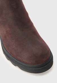 Detailní záběr na špičku hnědé semišové boty s černou gumovou podrážkou na bílém pozadí.