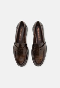 Mocasins de couro castanho com um acabamento brilhante, apresentando um design de biqueira cosida e um pequeno detalhe de correia na parte frontal. Forma suave e arredondada.