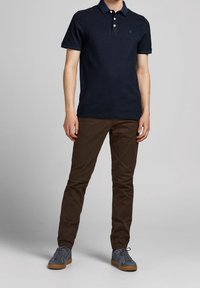 Marinblå polo-shirt med två knappar, korta ärmar och en liten logotyp på bröstet, tillsammans med bruna slim-fit chinos och grå sneakers.