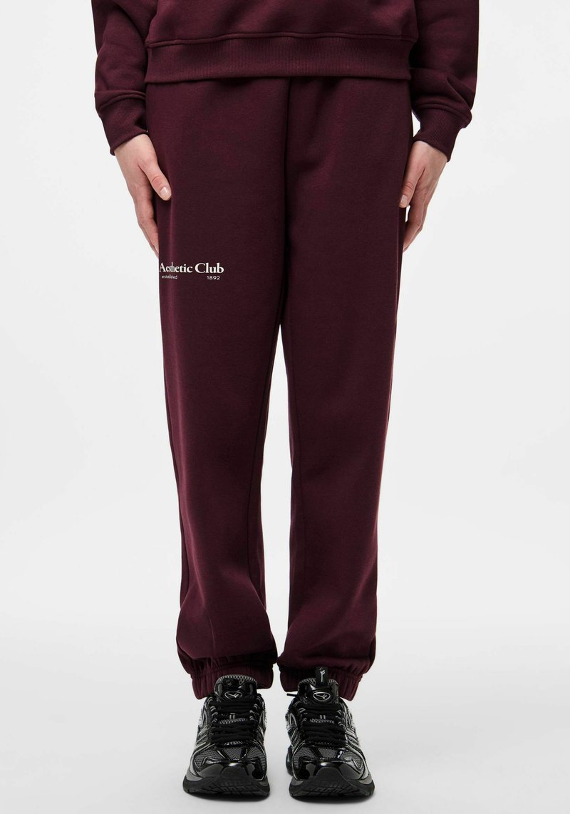 Bordo trenirke iz mehke tkanine z elastičnim pasom in manšetami. Spredaj je modno nameščen bel logotip.